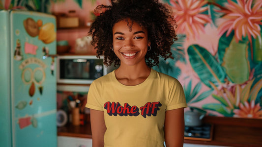 Smiling Black woman wearing a yellow t-shirt that reads “Woke AF” in bold red and black lettering, standing in a colorful retro kitchen with floral wallpaper and a pastel teal fridge. Represents joy, pride, and conscious self-expression.